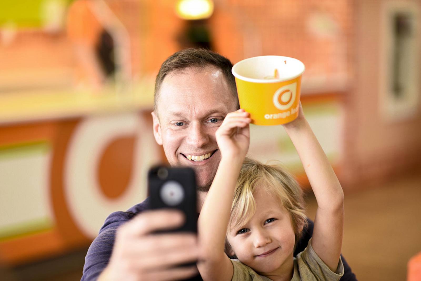 an Adult and Child Smile for a Selfie at Orange Leaf, Holding Frozen Yogurt During a Fun Outing, Colorful Decor in the Background. Commercial Photography – Amatucci Photography - Amatucci Photography An adult and child smile for a selfie at orange leaf holding frozen yogurt during a fun outing colorful decor in the background Commercial photography amatucci photography