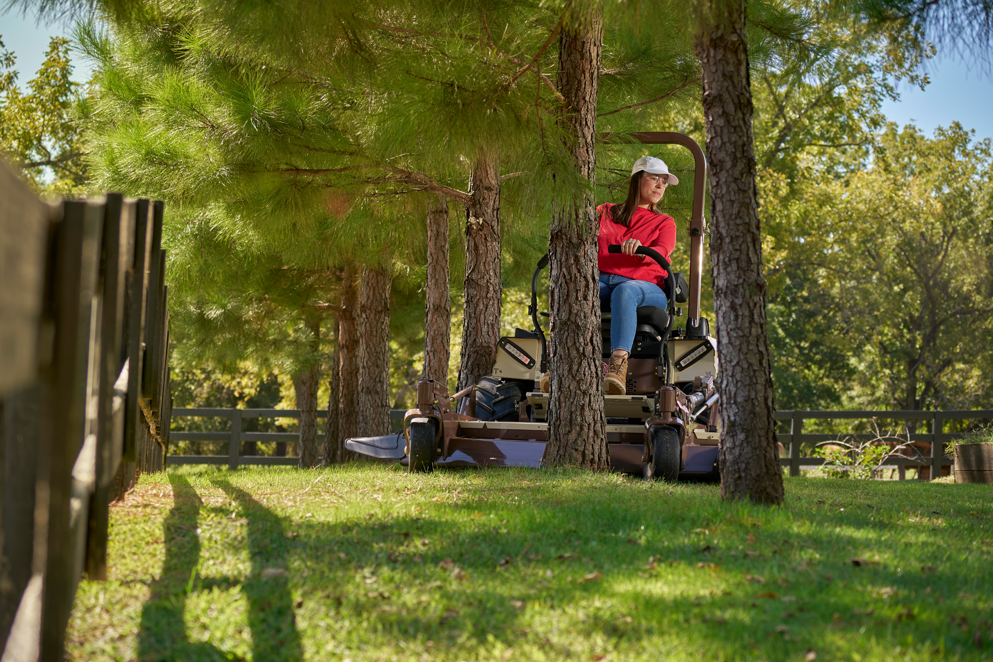 Someone in a White Cap and Red Shirt Mows a Sunlit Yard Under Pine Trees Near a Wooden Fence in a Park-like Setting. – Amatucci Photography - Amatucci Photography Someone in a white cap and red shirt mows a sunlit yard under pine trees near a wooden fence in a park like setting Commercial photography of a grasshopper lawnmower amatucci photography