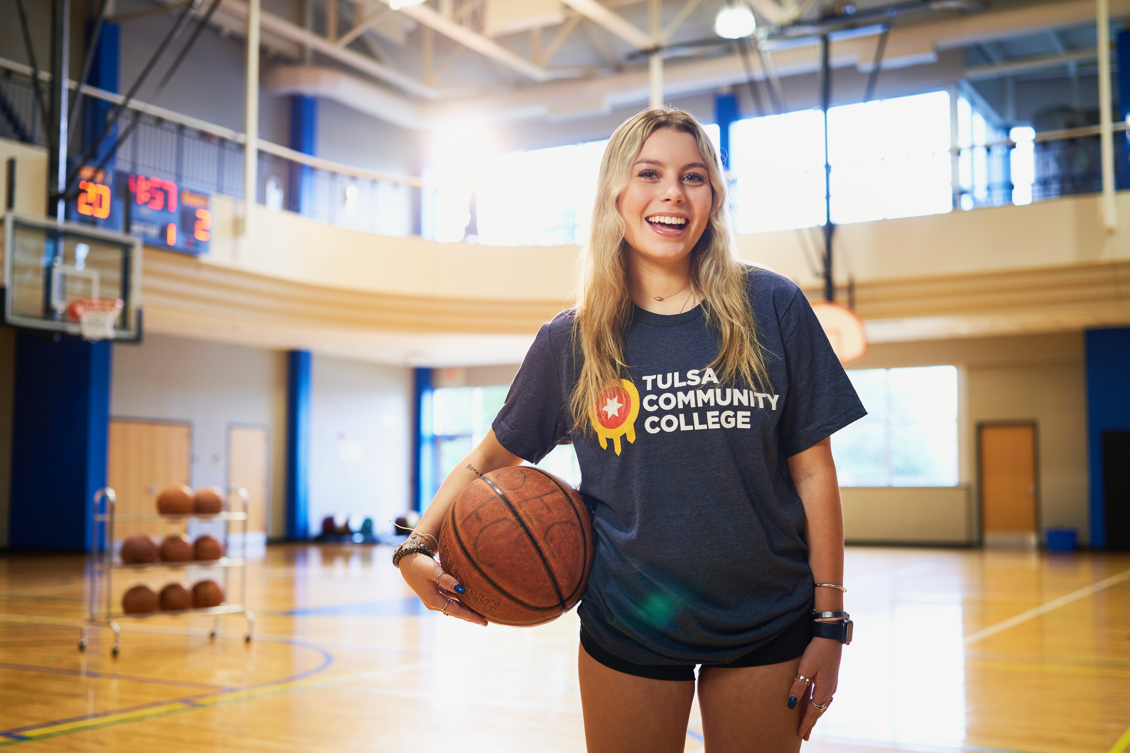 Smiling Tcc Student in College Gym Holds a Basketball During Sports Event, Bright Windows and Stacked Balls Behind Her. Commercial Photography – Amatucci Photography - Amatucci Photography Smiling tcc student in college gym holds a basketball during sports event bright windows and stacked balls behind her Commercial photography amatucci photography