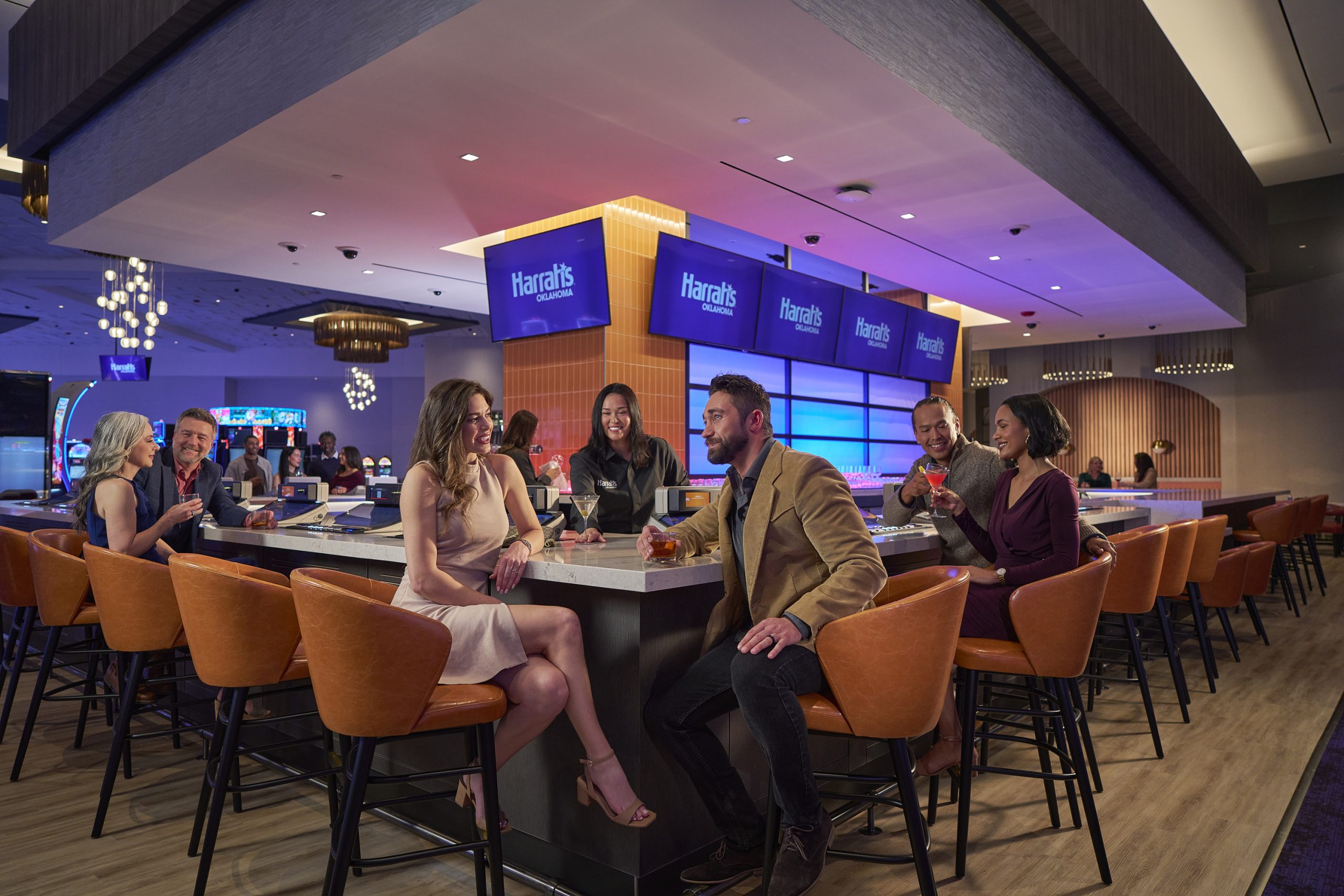 Friends Mingle with Drinks at Harrah's Oklahoma Casino Bar, Amid Orange Chairs, Slot Machines, Tvs, and Glowing Blue Signage. Commercial Photography – Amatucci Photography - Amatucci Photography Friends mingle with drinks at harrahs oklahoma casino bar amid orange chairs slot machines tvs and glowing blue signage Casino lifestyle campaign commercial photography amatucci photography